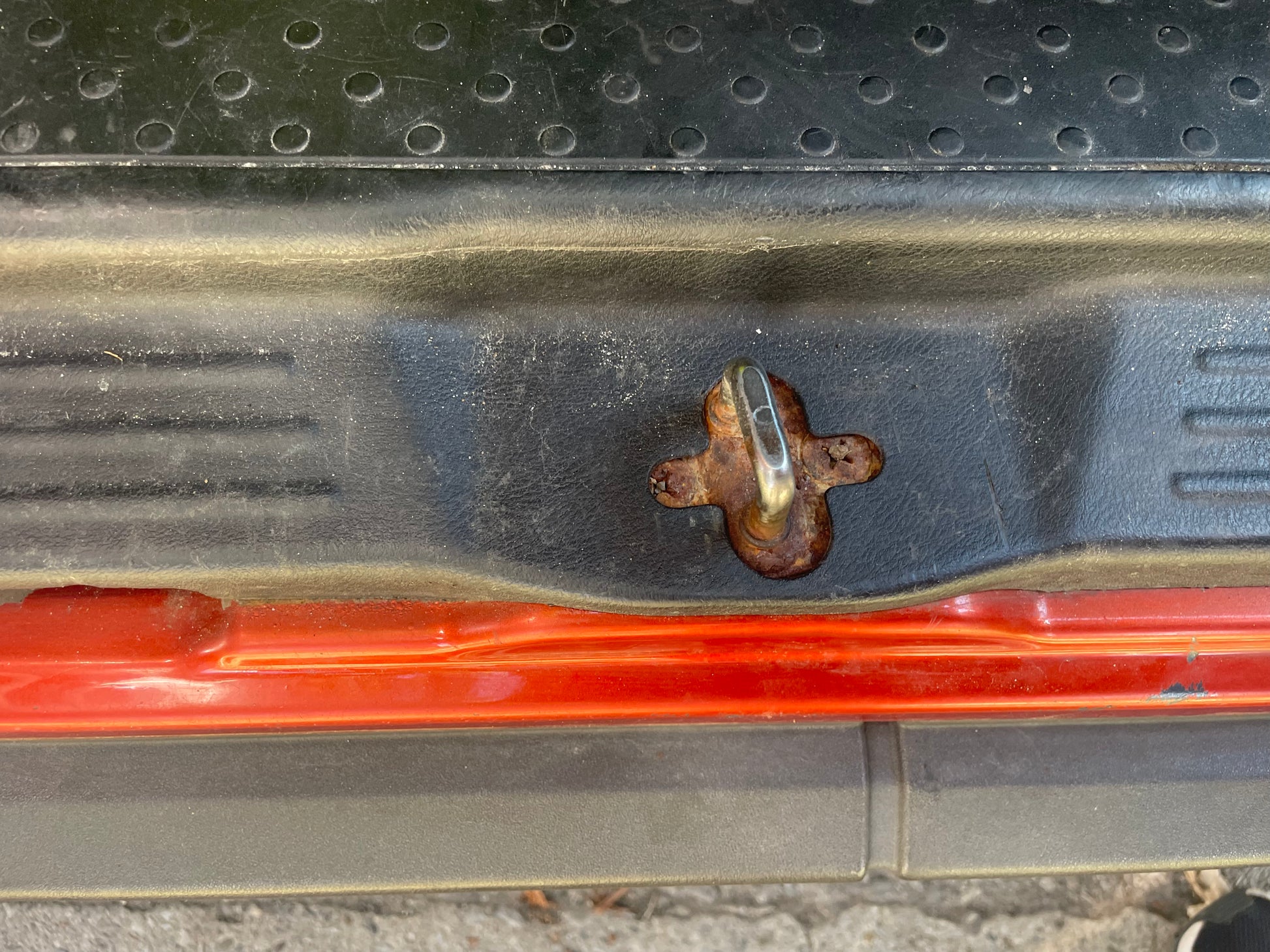 Close-up of a rusty car door latch on a black textured surface, with an orange door frame visible.