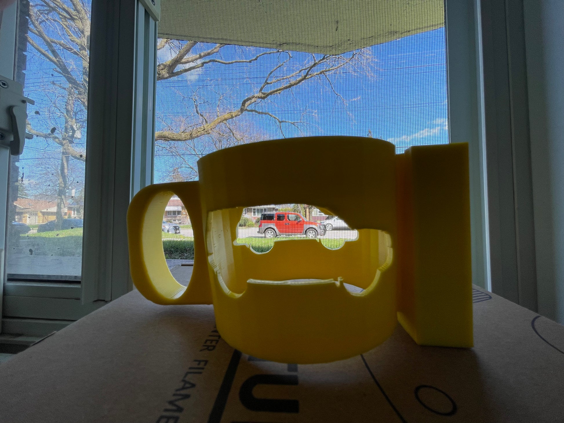 Yellow 3D printed mug with a cutout design on a box near a window.